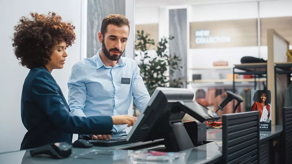 A woman and a man is next to the cashier in the retail shop, clearly shown that it is retail business and showing that they are solving workforce management issues